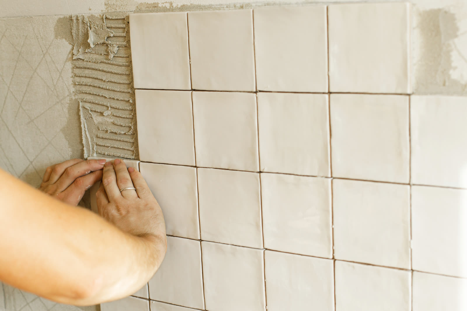 Hands pressing a square wall tile into fresh adhesive